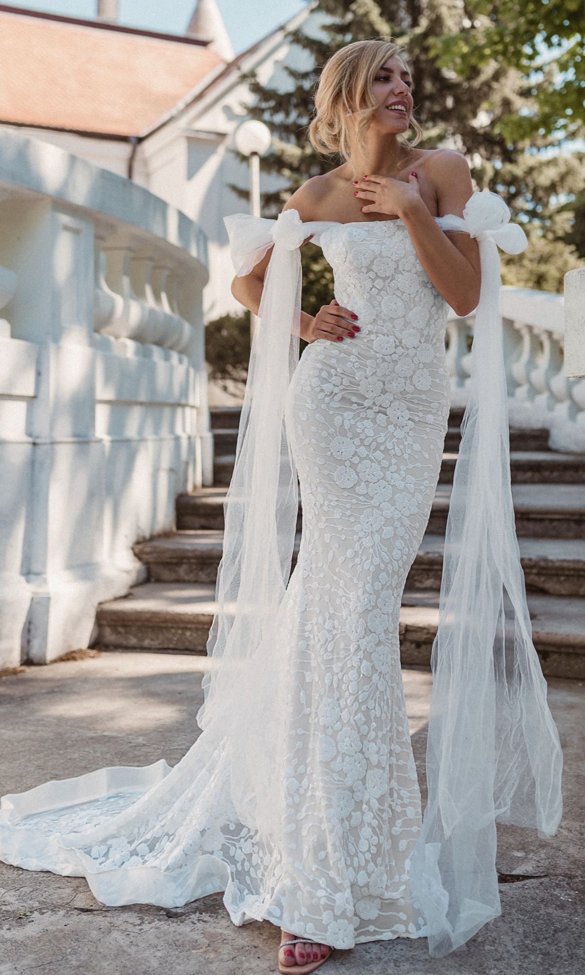 A woman in the Strapless White Wedding Gown: Tina Holly BB010, featuring a fitted mermaid silhouette and sheer train details, stands smiling to her left on outdoor stone steps.